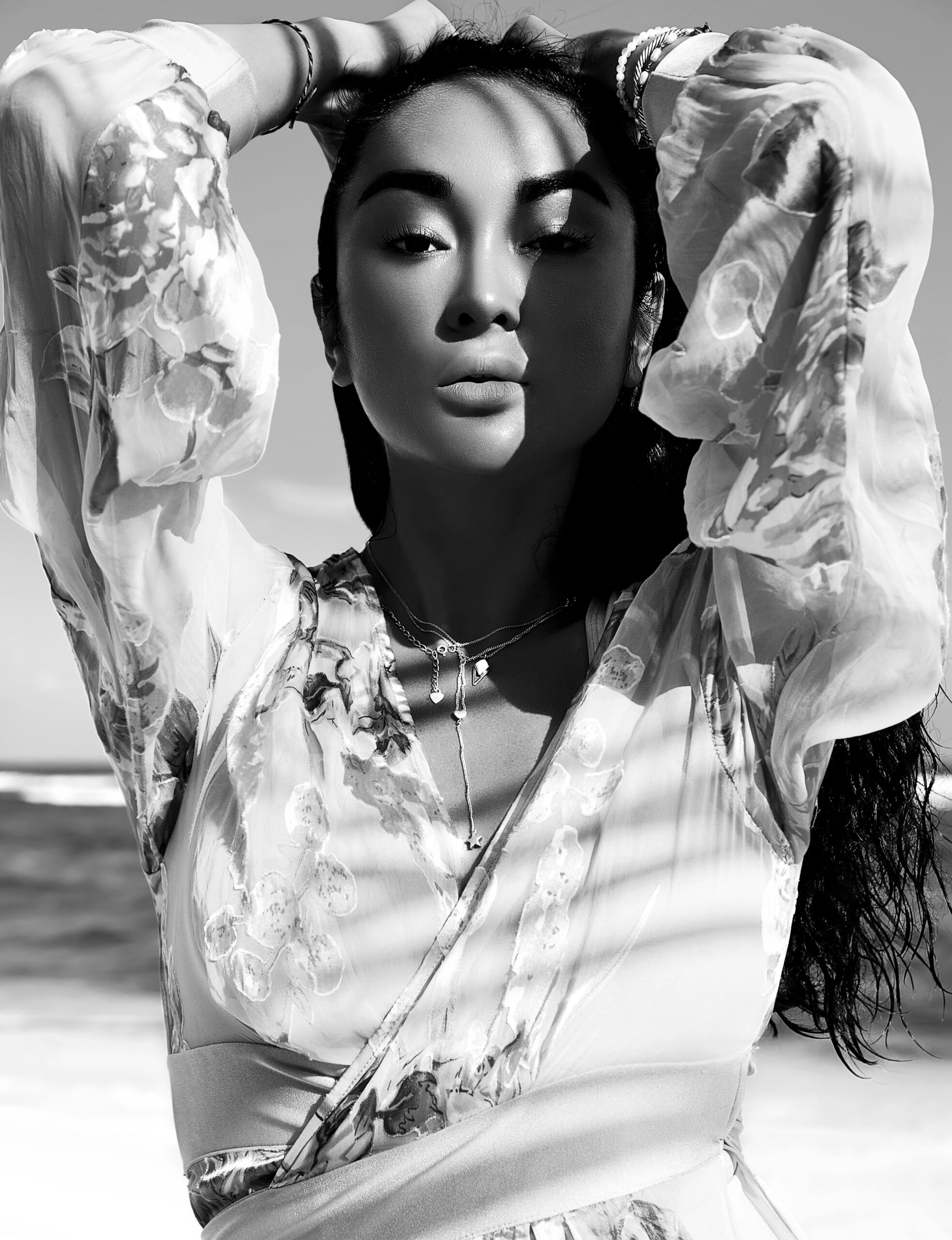 portrait of beautiful caucasian sunbathed woman model with dark long hair in beige flying dress posing on summer beach with white sand on blue sky and ocean background. touching her hair